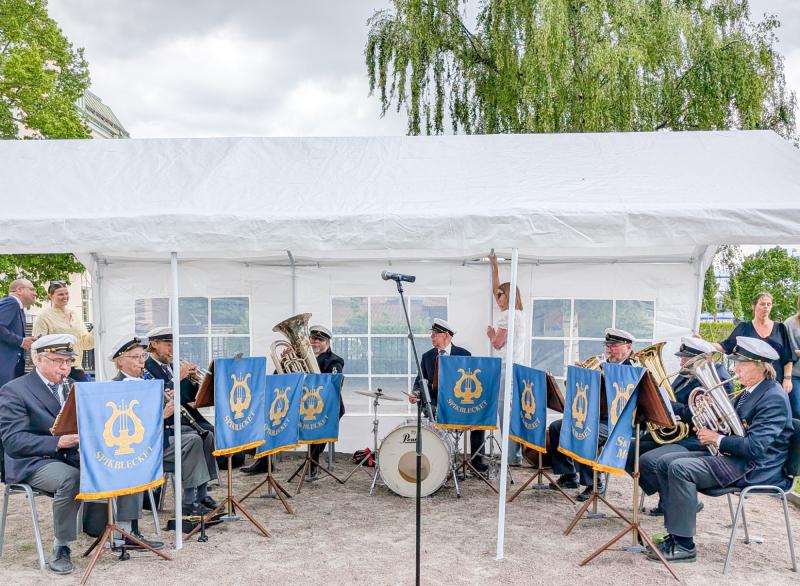 En blåsorkester uppträder under ett vitt tält. Uniformsklädda musiker spelar olika instrument och framför sig har de blå banderoller med en gyllene lyra och 