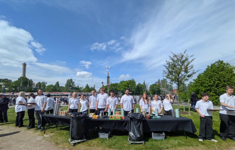 En grupp unga människor i vita skjortor och svarta byxor står bakom ett bord med mat och dryck, utomhus under en blå himmel med moln.