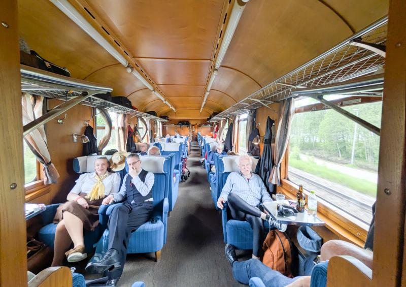 Interiörbild av en tågvagn med passagerare som sitter i blå sammetsstolar.