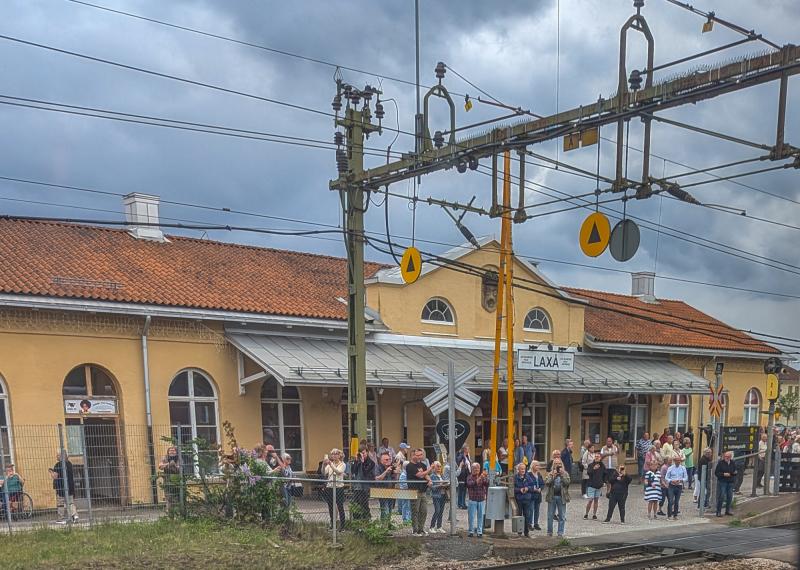 En folkmassa samlas utanför en gul tågstation med orange tegeltak, luftledningar och en molnig himmel.