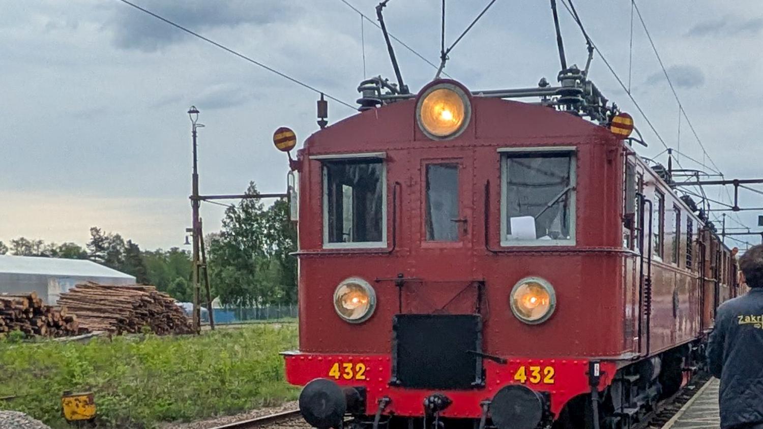 Ett rött elektriskt vintagelokomotiv med nummer 432 är på spåren under en mulen himmel. Timmer ligger staplat till vänster.