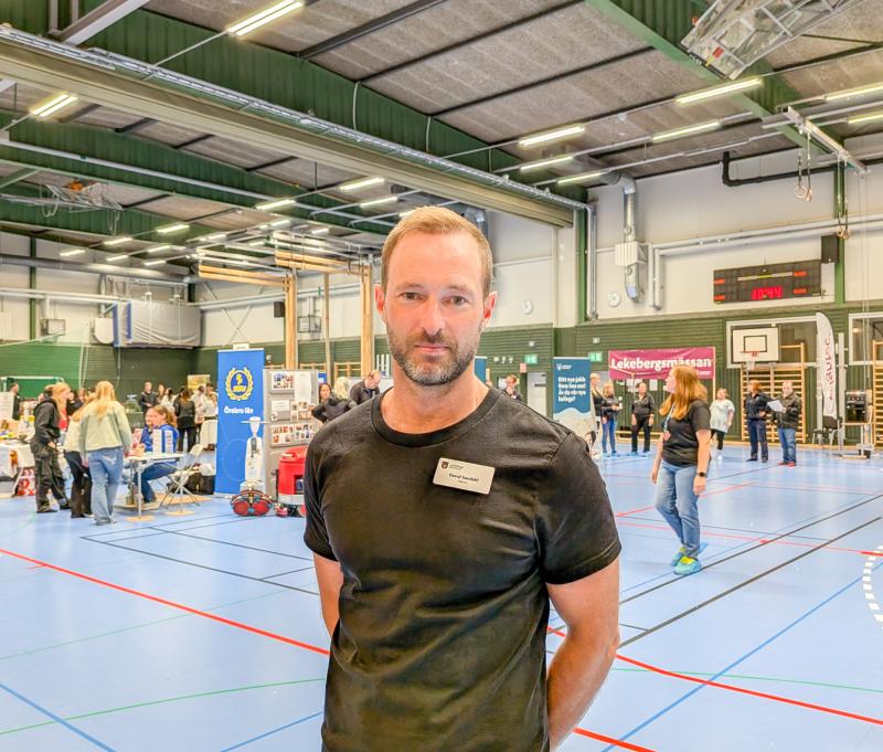 En man med en namnskylt står i en gymnastiksal på ett evenemang med informationsbås och människor i bakgrunden.