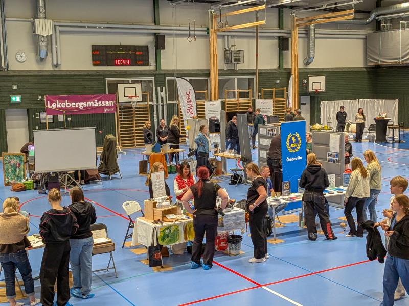 Inomhusmässa med flera montrar och människor som interagerar i en gymnastiksal. På utställningsborden finns skyltar, broschyrer och utställningsföremål. Basketkorgar och en resultattavla är synliga.