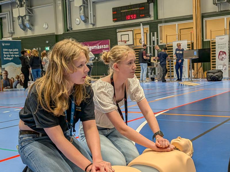 Två ungdomar övar bröstkompressioner på en träningsdocka för HLR i en gymnastiksal under ett offentligt evenemang.