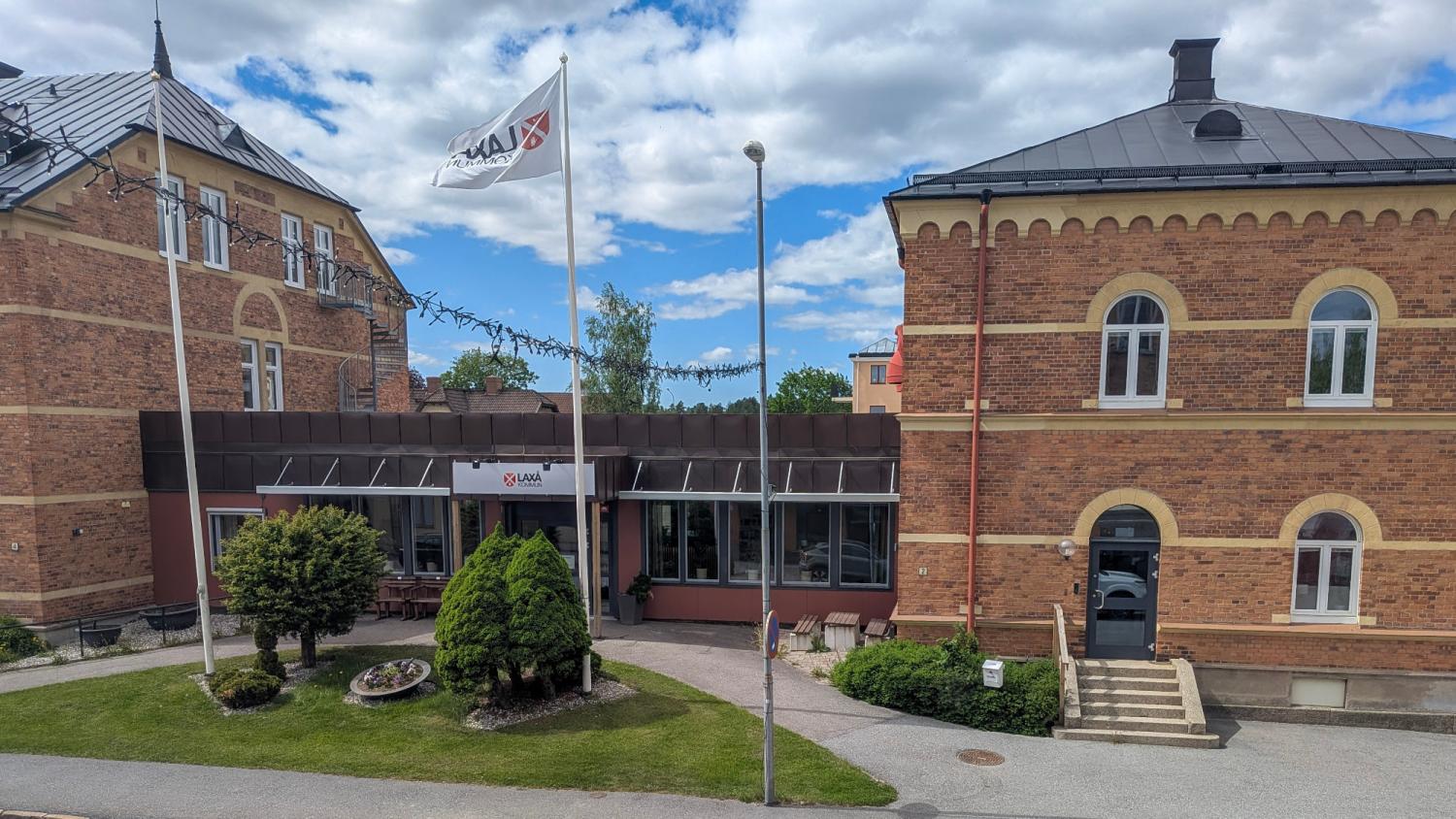 En tegelbyggnad med svart plåttak, en flaggstång med vit flagga och en huvudentré omgiven av buskar och träd under en delvis molnig himmel.