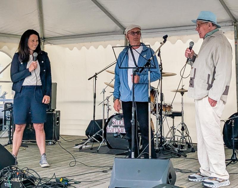 Tre personer står på en utomhusscen under ett tält, var och en med en mikrofon i handen, medan musikinstrument och utrustning syns i bakgrunden.