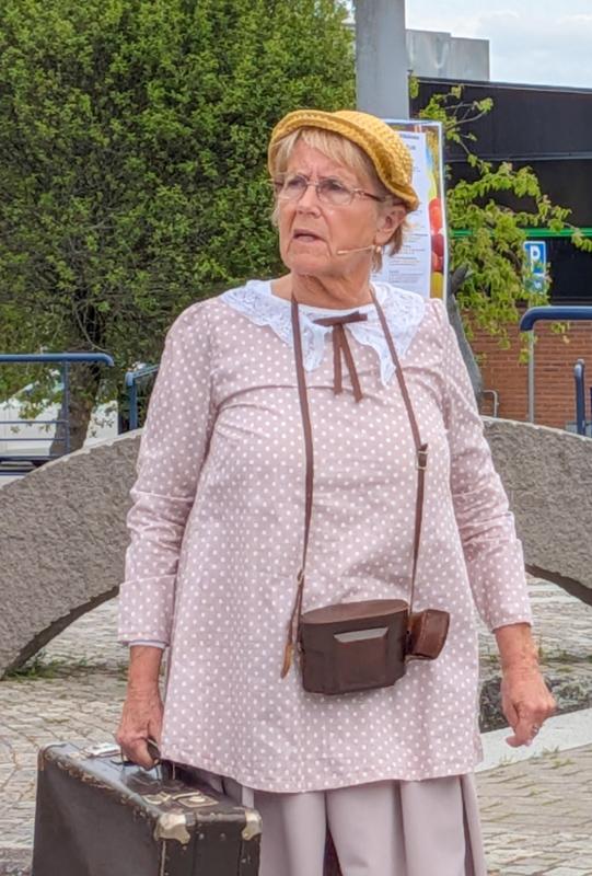 En äldre kvinna i en prickig klänning står utomhus med en resväska i handen. Hon bär en gul hatt, glasögon och ett mikrofonheadset, med träd och en byggnad i bakgrunden.