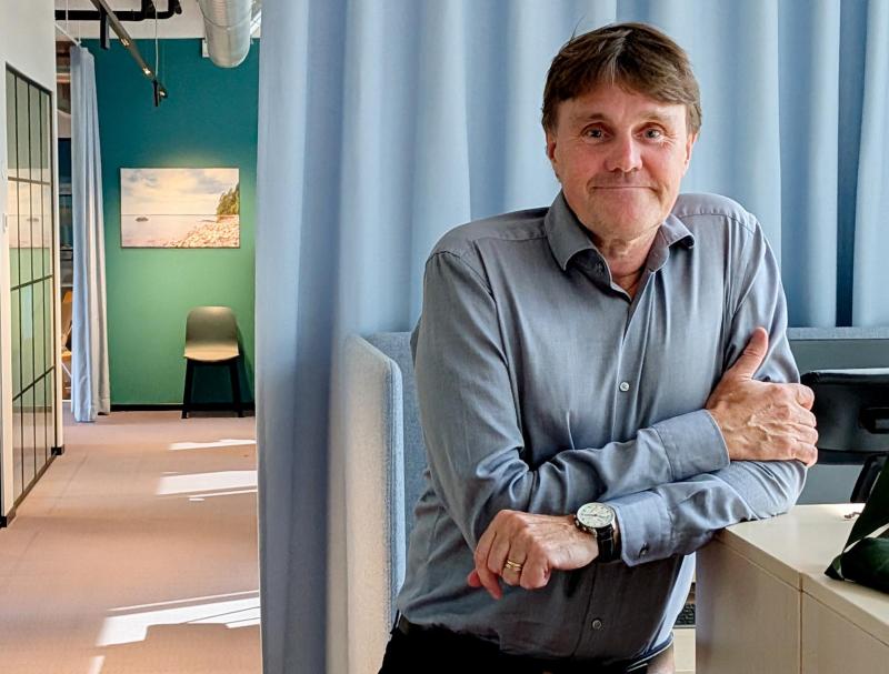 En man i en grå skjorta står vid ett skrivbord i ett modernt kontor där man i bakgrunden kan se blå gardiner, en stol och en tavla.