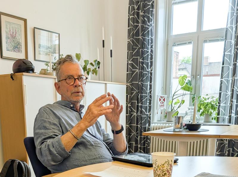 En äldre man med glasögon sitter vid ett skrivbord på ett kontor och gestikulerar med sina händer. I bakgrunden syns papper, en kaffekopp och växter.