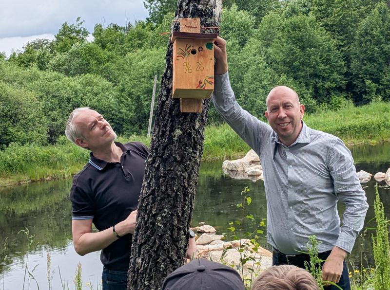 Två män står vid en damm; den ene håller i en dekorerad fågelholk av trä som är fäst vid ett träd, omgivna av grönska.