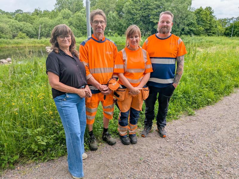 Fyra vuxna står på en grusgång nära grönskan; tre av dem bär orange arbetskläder och en är klädd i fritidskläder.