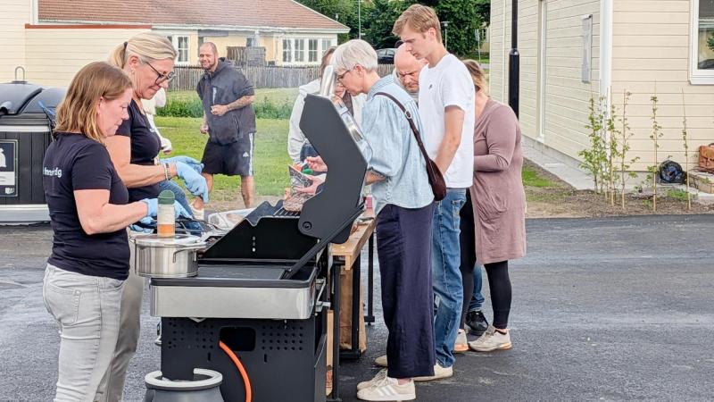 En grupp människor står runt en utomhusgrill och lagar och serverar mat på en stenlagd yta intill ljusa byggnader.