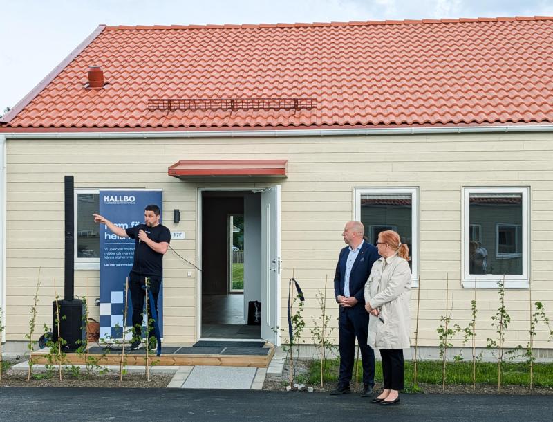 Tre personer står utanför ett litet beige hus med rött tegeltak; en man talar i en mikrofon medan de andra två lyssnar.
