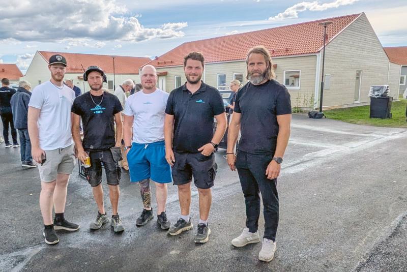Fem män står utomhus på en stenlagd yta framför envåningshus med röda tak under en delvis molnig himmel.
