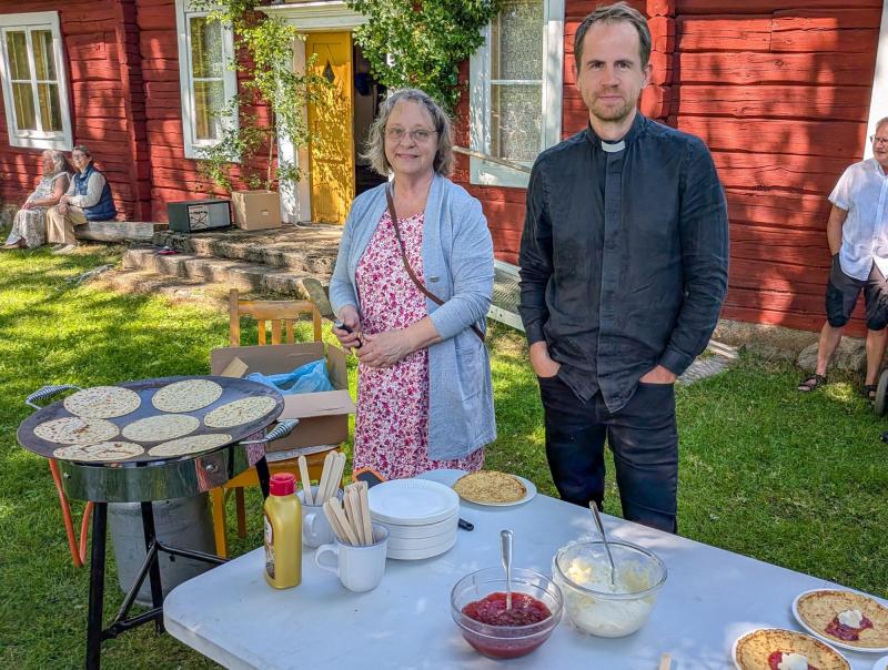 Två personer står bakom ett bord med tallrikar, kryddor, sylt och grädde, bredvid en stekhäll med pannkakor. I bakgrunden syns en röd träbyggnad och andra människor.