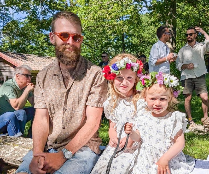 En man sitter på en bänk bredvid två unga flickor som bär blomsterkransar och matchande klänningar vid en utomhussammankomst. I bakgrunden syns fyra andra vuxna.