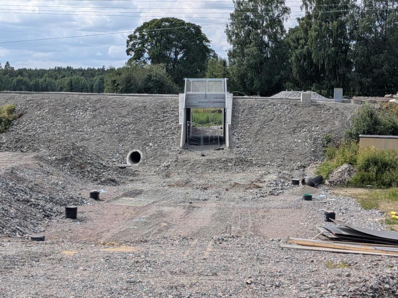 En grusbelagd byggarbetsplats med en kulvert eller undergång med en inhägnad ingång, ett dräneringsrör och utspridda byggmaterial. I bakgrunden syns träd och kraftledningar.