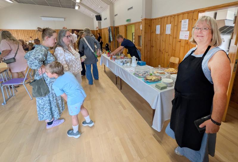 Folk står i kö vid ett bord med mat och dryck i en föreningslokal. En kvinna i förkläde står i förgrunden och håller i ett svart föremål, medan andra väntar på att bli serverade.