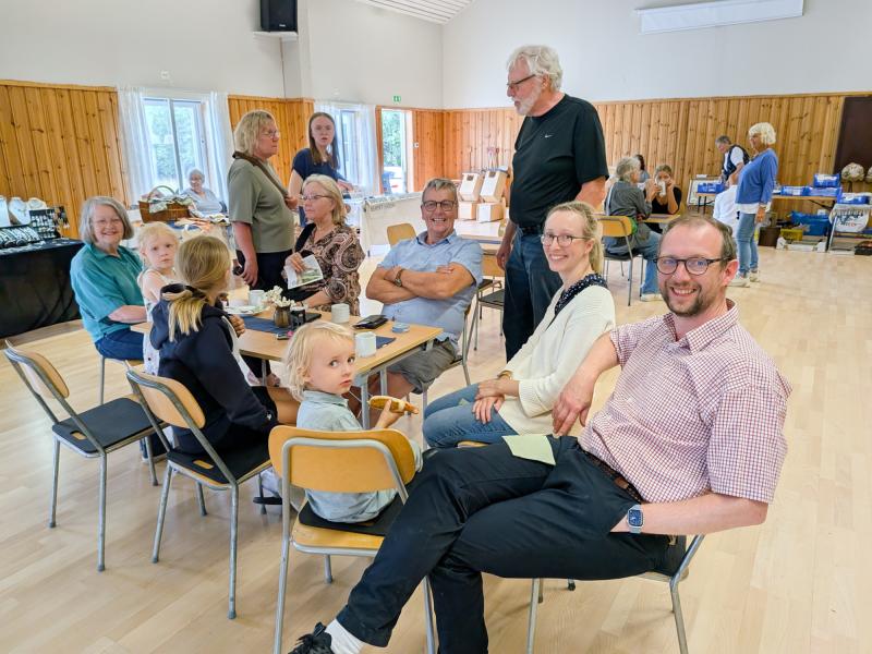 En grupp vuxna och barn sitter och står runt borden i en samlingslokal och deltar i samtal och aktiviteter.