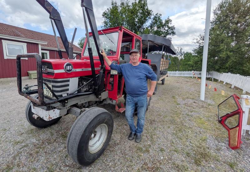 En man i blå skjorta och keps står bredvid en röd traktor som står parkerad på en grusuppfart, med en vagn kopplad bakom sig och träd i bakgrunden.