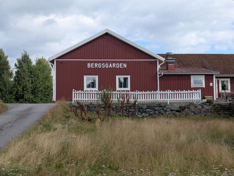 En röd träbyggnad med vita lister och skylten ”Bergsgården”, omgiven av en stenmur, ett vitt staket, gräs och träd under en molnig himmel.