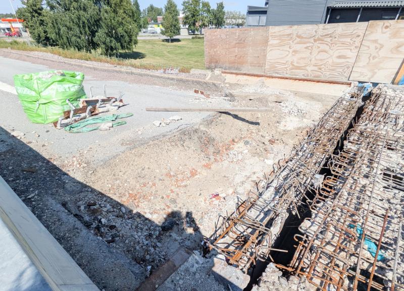 En byggarbetsplats med synliga armeringsjärn, byggavfall och en grön säck med material en solig dag; ett trästaket och en gräsbevuxen yta i bakgrunden.