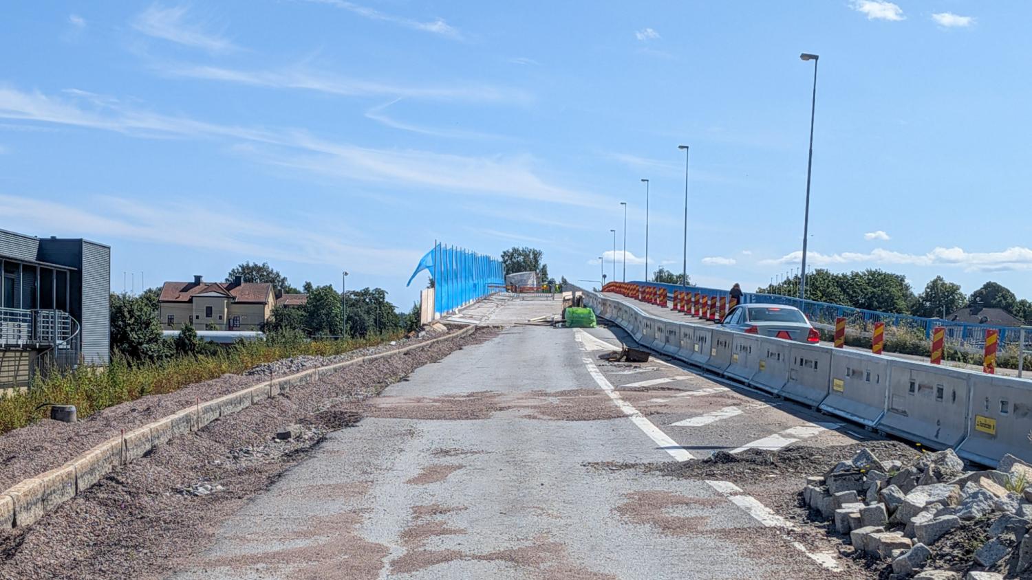 En delvis färdigställd väg med byggstaket, byggavfall och ett fordon i fjärran. Blå stängsel kantar den ofärdiga sträckan under en klar himmel.