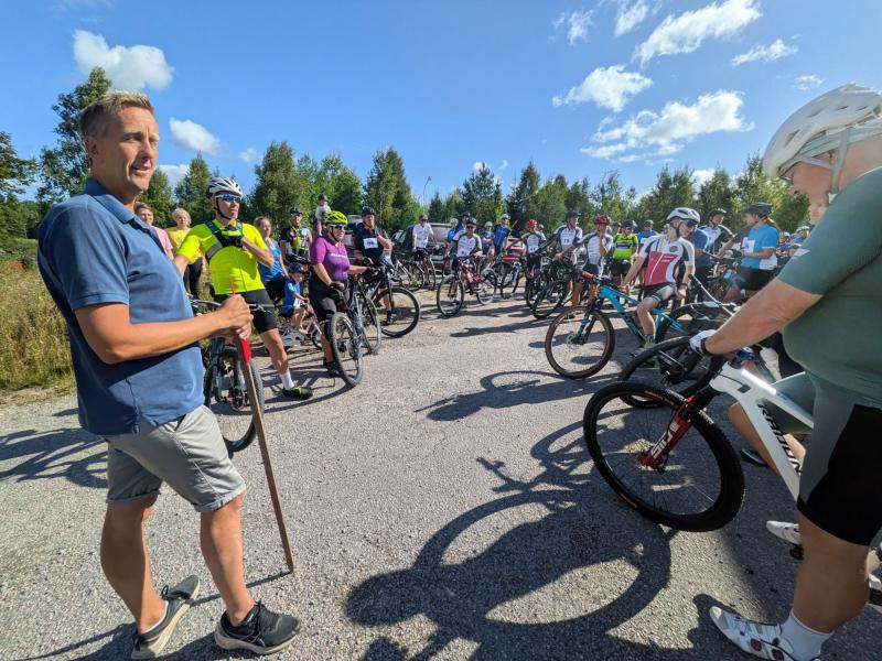 En grupp cyklister har samlats utomhus en solig dag och förbereder sig för en cykeltur, medan en man i vardagskläder står med en käpp i förgrunden.