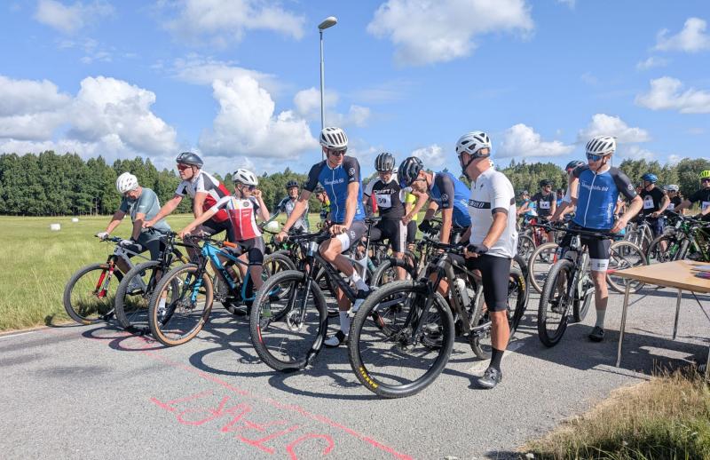 Cyklister med hjälmar och cykeltröjor ställer sig upp vid startlinjen en solig dag och gör sig redo för ett lopp på en landsväg.