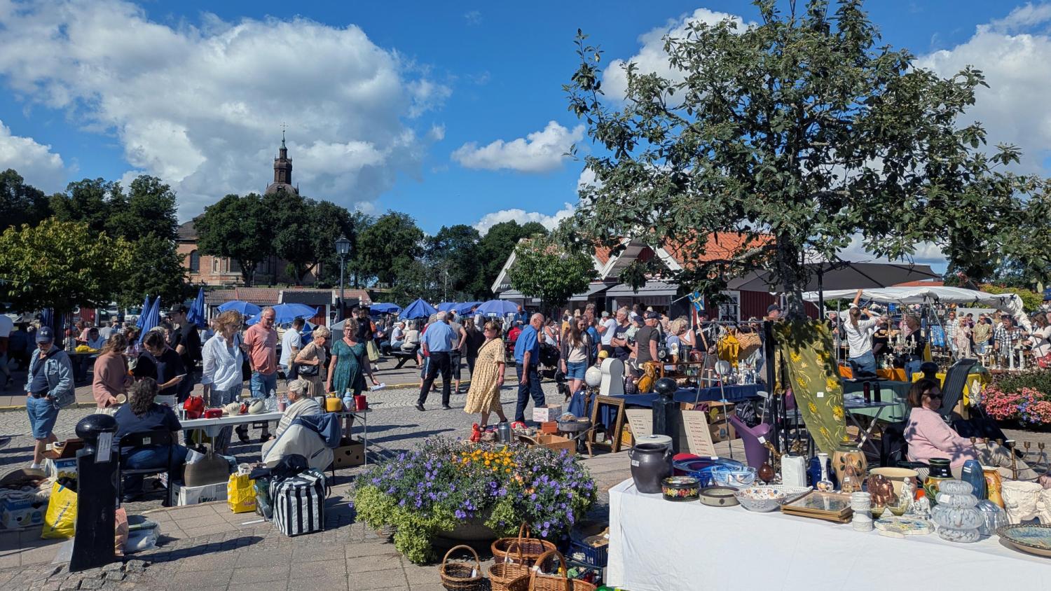 Folk strosar bland stånden och borden med olika varor på en utomhusmarknad en solig dag, med träd, blommor och en kyrka synliga i bakgrunden.