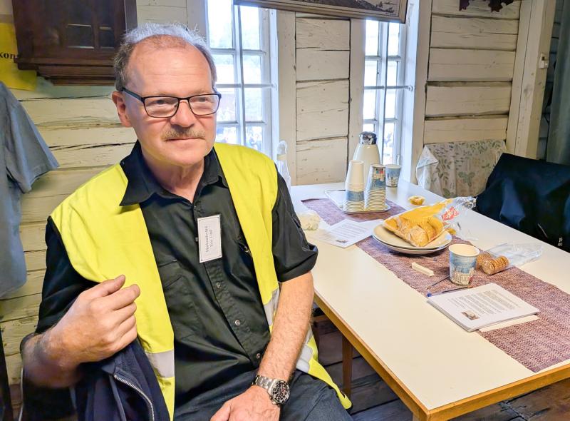 En man med glasögon och mustasch sitter vid ett bord med bröd, bakverk, tidningar och en kaffeservis i ett rustikt rum med vita träväggar och fönster.
