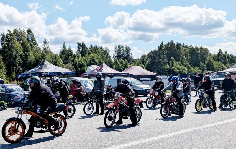 En grupp människor på mopeder och motorcyklar samlas en solig dag, med tält, bilar och träd synliga i bakgrunden.