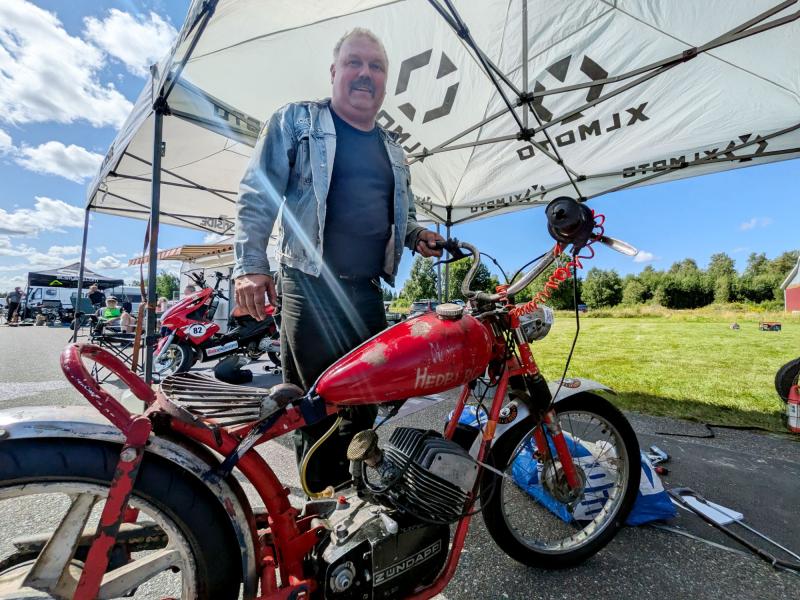 En man som står bredvid en röd Henderson-motorcykel i retrostil under ett tält vid ett utomhusevenemang, med en gräsbevuxen yta och blå himmel i bakgrunden.