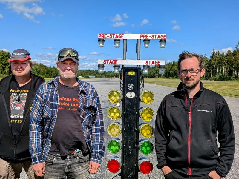 Tre män står bredvid en startlampa för dragracing på en racerbana, med träd och blå himmel i bakgrunden.