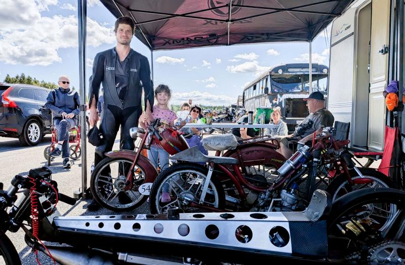 En man i racingkläder och ett barn står under ett tält bredvid klassiska motorcyklar vid ett utomhusevenemang, med människor och fordon i bakgrunden.