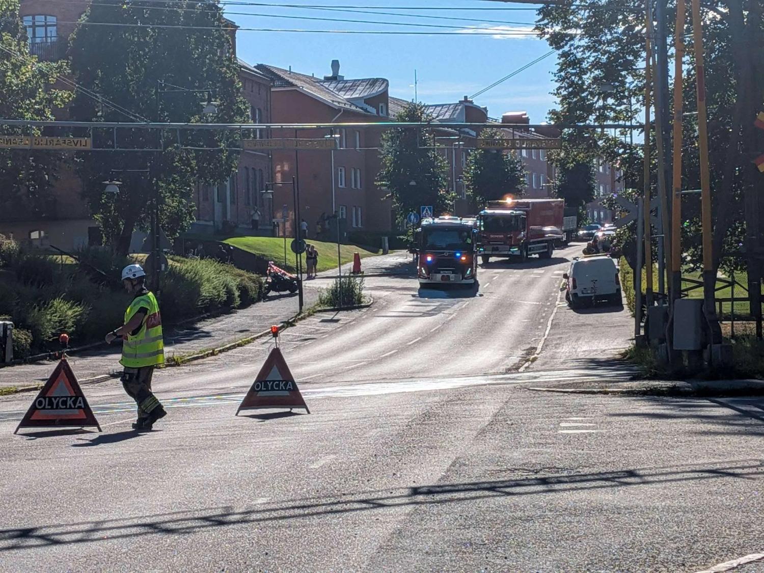 Brandbilar och räddningspersonal spärrar av en gata med varningsskyltar med texten ”Olycka” (olycka) en solig dag i ett bostadsområde.