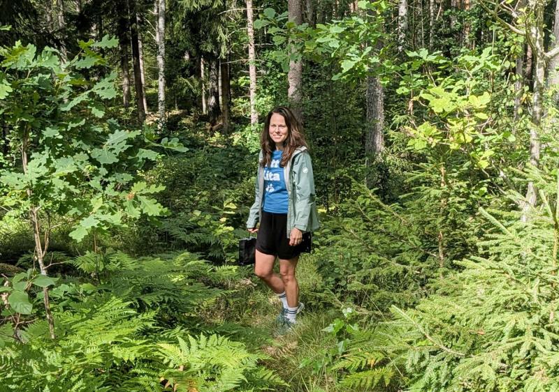 En kvinna står på en smal stig omgiven av gröna träd och ormbunkar i en solbelyst skog och ler mot kameran.