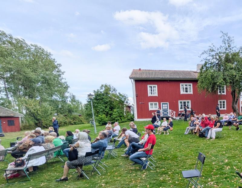 En grupp människor sitter på stolar utomhus framför en röd träbyggnad och deltar i ett evenemang på en gräsmatta under en delvis molnig himmel.