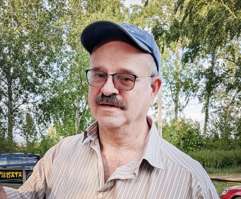 En man med glasögon, mustasch och en blå keps står utomhus framför gröna träd. Han har på sig en randig skjorta och tittar mot kameran.