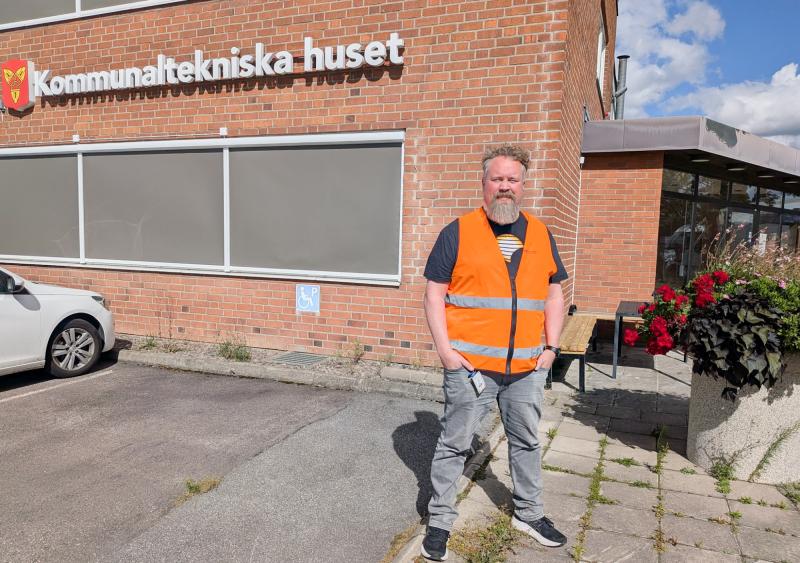 En man i en knallorange säkerhetsväst står utanför en tegelbyggnad med skylten ”Kommunaltekniska huset” en solig dag.