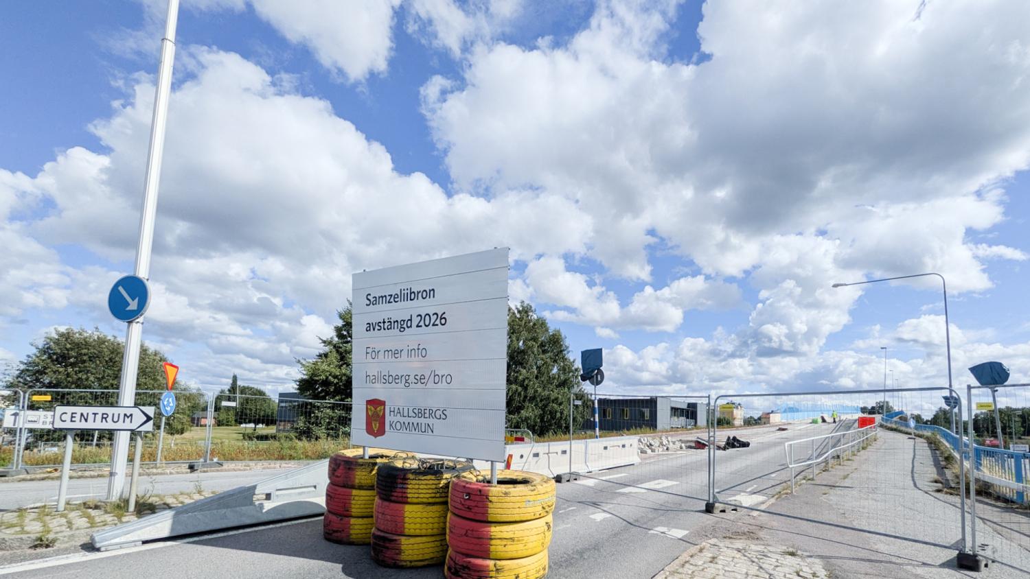 En väg är avstängd med avspärrningar och en skylt med texten ”Sannaledsbron färdig 2026” på svenska. Himlen är delvis molnig och i bakgrunden syns byggarbete.
