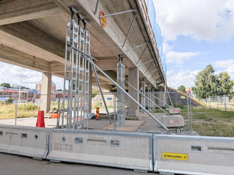 En betongbro stöds av tillfälliga metallstöd och avspärrningar. Byggskyltar och stängsel begränsar tillträdet under konstruktionen.