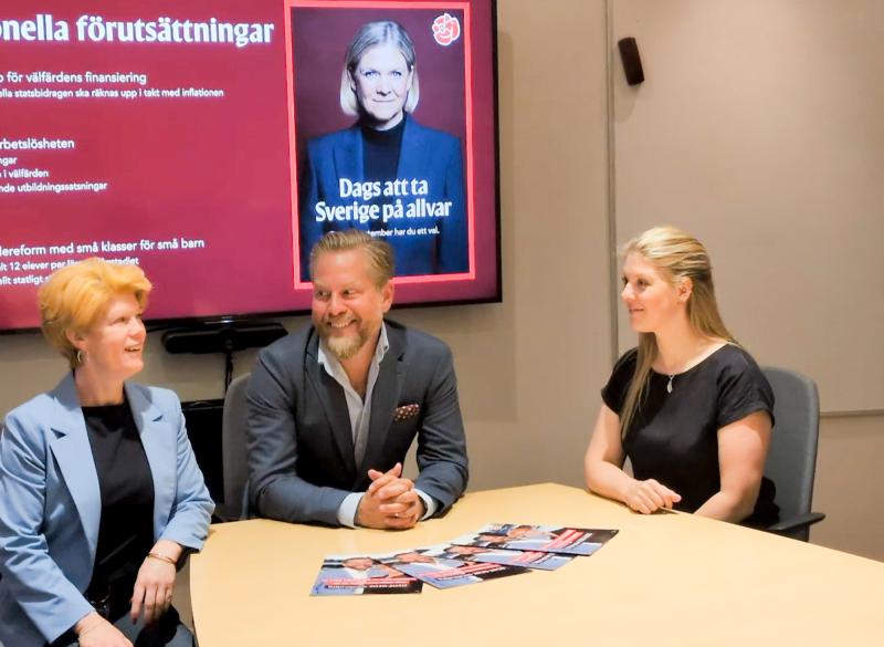 Tre personer sitter vid ett konferensbord med broschyrer och pratar. På en digital skärm bakom dem visas en politisk reklamfilm med en bild på en kvinna och svensk text.