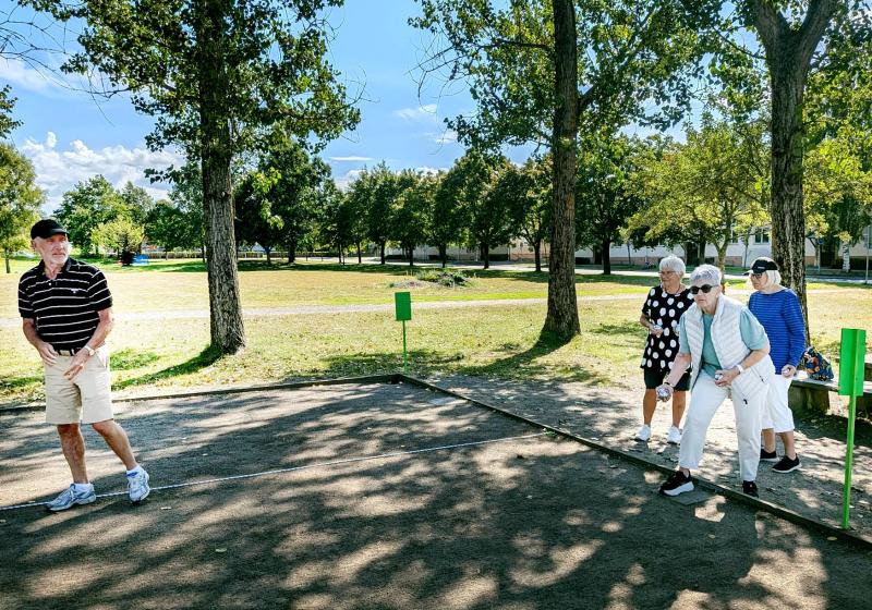 Fyra äldre personer spelar ett spel utomhus en solig dag, stående på en grusplan omgiven av gräs och träd.