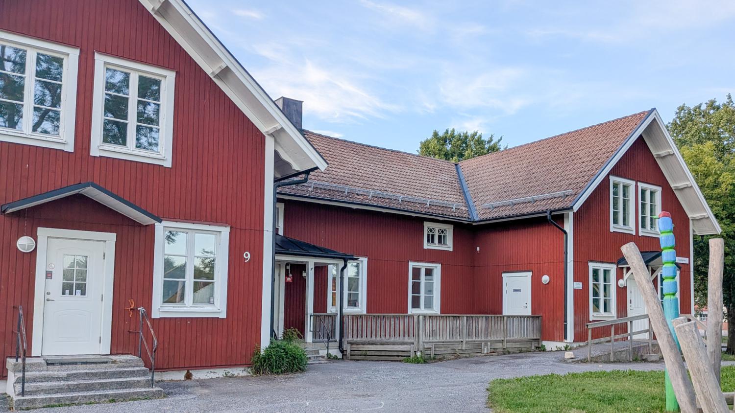 En röd träbyggnad med vita lister, flera fönster, ett sadeltak och en liten ramp som leder till ingången. I bakgrunden syns träd och en färgglad stolpe.