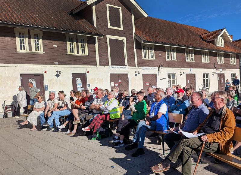 En grupp människor sitter på stolar utanför en byggnad med brunt fasadpaneler och rött tak och lyssnar på ett evenemang eller en presentation en solig dag.