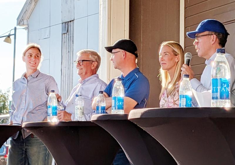 Fem personer sitter bakom ett bord med mikrofoner och vattenflaskor och deltar i en paneldiskussion utomhus en solig dag.