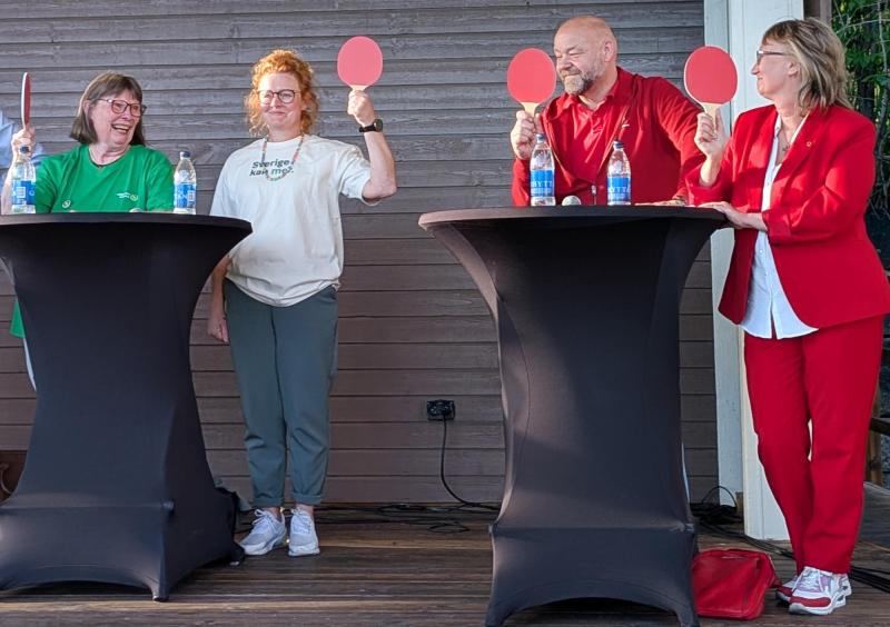 Fyra personer står vid cocktailbord och håller var och en upp en röd paddel; det ser ut som om de deltar i en paneldiskussion eller ett spel. På borden står vattenflaskor och mikrofoner.