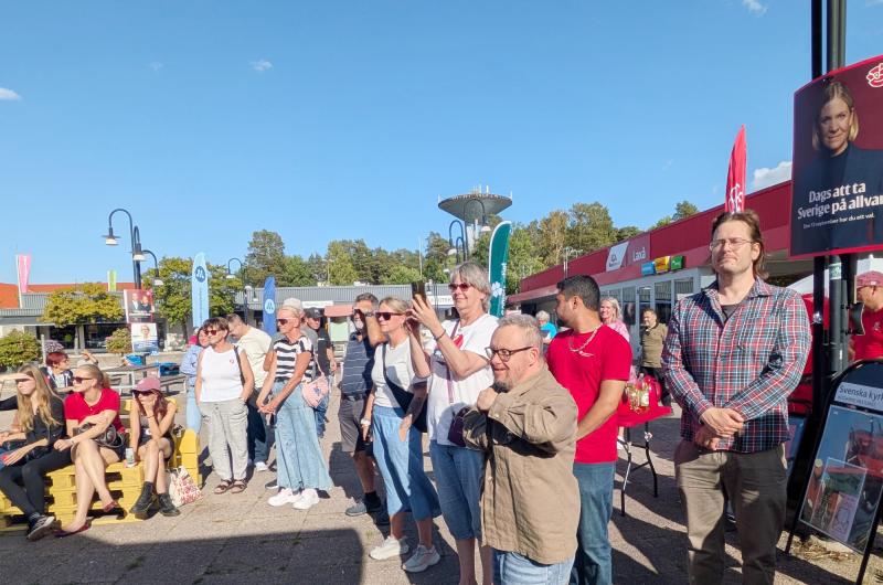 En grupp människor står utomhus vid ett evenemang där banderoller, skyltar och montrar syns under en klar himmel.