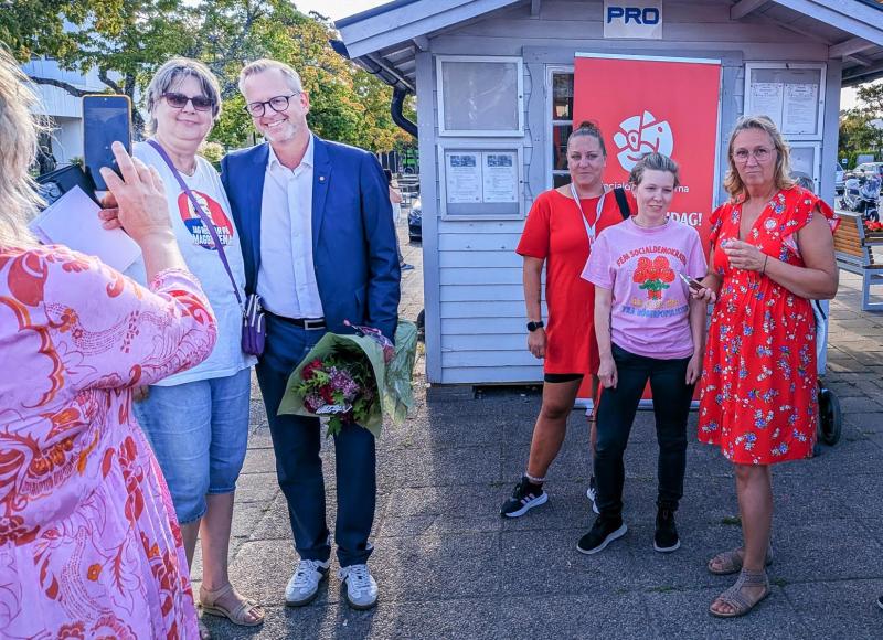 En grupp på sex personer poserar för ett foto utanför en liten byggnad; en person håller blommor och en annan tar en bild med en mobil.